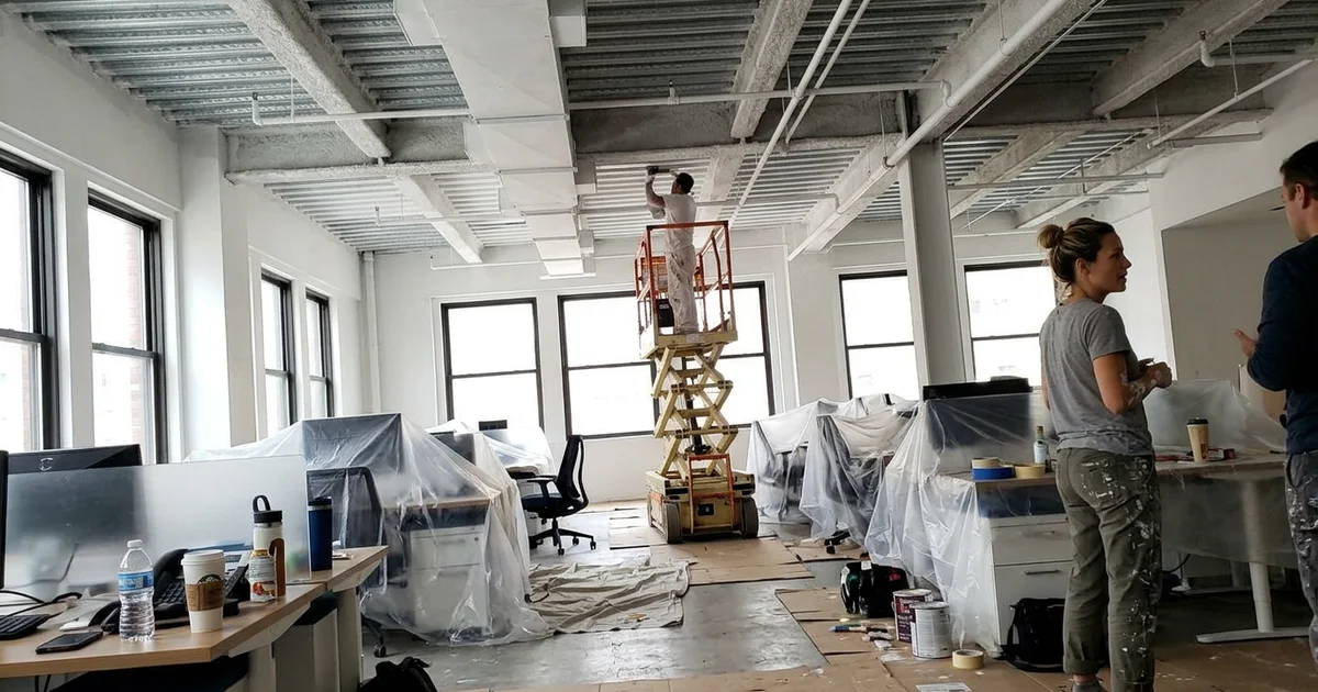Open-plan office with a high exposed ceiling and a scissor lift set up for painting