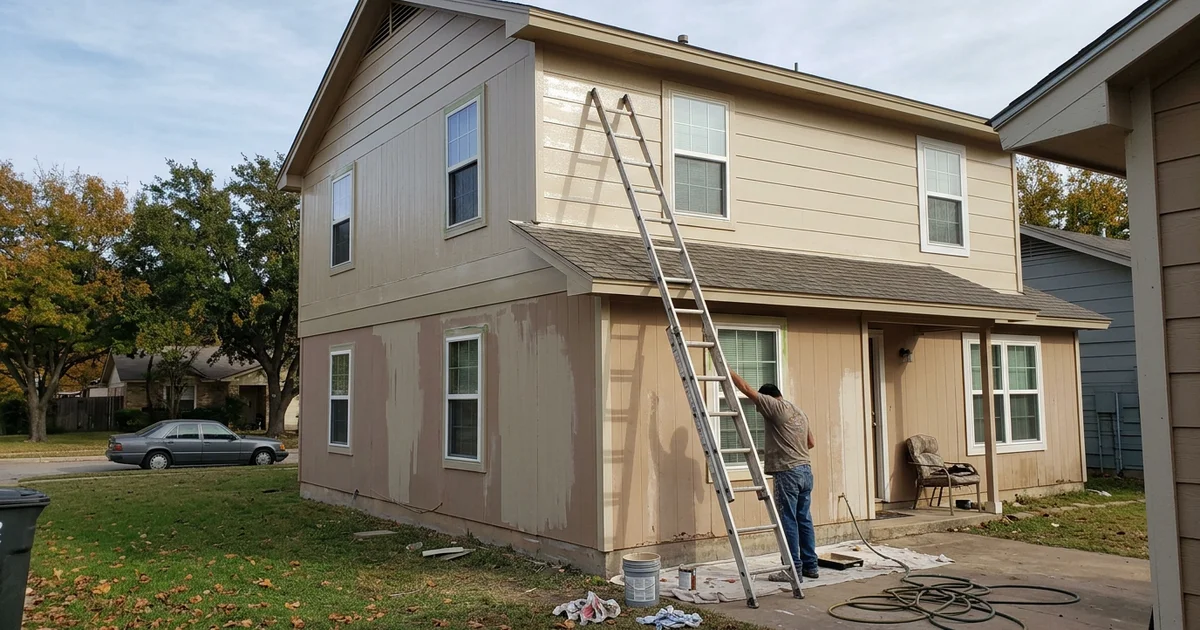 Two-story fiber cement house mid-repaint with a ladder against the wall on a fall morning